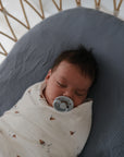 A newborn sleeps peacefully with a pacifier, wrapped in a white sailboat-printed blanket, resting on the mushie Mini Muslin Crib Sheet—made from 100% cotton—inside a wicker crib.
