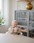A smiling toddler in a high chair holds a mug at a round table, enjoying their mushie Deluxe Pink Flowers/Blush Feeding Kit. Behind them is a glass cabinet with glassware, a window with plants, and a large gold vase of dried flowers.