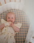 A baby in a cream dress with colorful dots smiles in a crib with a checkered mattress, holding the mushie Rainbow Teething Mitten.