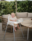 A smiling toddler sits outdoors in a white high chair on a wooden deck with the mushie Snack Cup in front of them. A wicker chair, round table, and green leafy bushes are visible in the background.