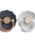 The FRIGG Daisy Natural Rubber Pacifier 2-Pack includes two daisy-shaped pacifiers with scalloped edges, circular bases, natural rubber latex nipples, and the FRIGG logo; one is dark gray/brown, the other white/light brown.