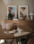 A wooden dining table with candles, mushie Bluey x Mushie Silicone Baby Bibs, and mugs sits in a cozy room. Behind it, a sideboard holds houseplants, lamps, and abstract art prints on the wall for a warm and inviting feel.