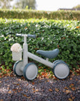Pale green mushie Mini Toddler Bike with basket of white flowers on gravel by green hedge.