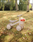 Mushie Mini Toddler Bike with floral basket on a grassy, leafy lawn outdoors.