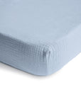 Close-up of the corner of a mushie Mini Muslin Crib Sheet, featuring textured light blue 100% cotton muslin with a soft, quilted look and breathable feel, shown fitted on a mattress against a white background.