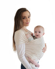A woman with long brown hair holds a baby snugly in a mushie Baby Wrap against a white background. Both face forward, and the baby looks at the camera, comfortable in the breathable carrier.