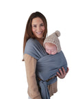 A smiling woman holds a sleeping baby in the mushie Baby Wrap, made from light blue organic cotton. The breathable wrap offers cozy, stylish comfort for baby wearing. A knitted pom-pom hat completes the look against the white background.
