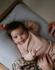 A baby in a light pink outfit lies on a cushioned changing pad, smiling up. An adult hand holds a silicone baby toothbrush, and the FRIGG Moon Natural Rubber Pacifier from FRIGG rests nearby among other baby essentials.