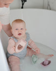 A baby in a patterned swimsuit sits in a bathtub, smiling and holding a non-toxic mushie Bath Boats toy, while an adult gently supports them. Two more stackable mushie Bath Boats float nearby, making bath time extra fun.