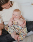 A light-skinned baby with blue eyes smiles and holds a teething toy while wrapped in a mushie Organic Cotton Muslin Swaddle Blanket, wearing a colorful dress on an adult’s lap, sitting on a bed with white pillows.