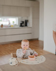 A baby sits in a high chair at a bright, modern kitchen table, happily eating from a mushie Snack Cup while an adult smiles nearby—perfect for toddlers learning to feed themselves.