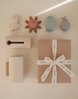 The mushie Sealife Bath Essentials Kit, including pastel baby toys, a mug, tissue holder, and ribbon-tied gift box, sits on a sunlit surface—perfect for gifting bath time essentials.