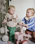 Two toddlers ride mushie Mini Toddler Bikes indoors; one drinks from a cup while the other watches.