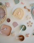 A pastel table setting with divided plates, cups, bowls, utensils, silicone toys, a rainbow cake, and FRIGG Daisy Natural Rubber Pacifier 2-Pack by FRIGG—all in soft, muted tones—arranged neatly on a white surface.
