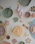 A pastel table setting includes FRIGG Butterfly Anatomical Silicone Pacifier 2-Pack (6-18 Months), children's plates, cups, utensils, toy blocks, candles, and a rainbow-shaped cake—all arranged in a circle on a white surface.