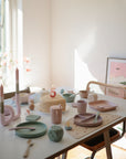 A sunlit table set for a child's 5th birthday features pastel dishes, FRIGG Butterfly Anatomical Silicone Pacifiers (6-18 months), a cake with a number 5 candle, pink candles, party decorations, a chair, and a large pink painting in the background.