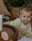 A smiling, light-haired baby in a cream knit sweater lies on a wooden floor with a FRIGG Silicone Pacifier 6-Pack (by FRIGG) in their mouth; soft toys, household items, and a wicker basket with greenery are nearby.