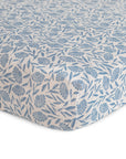 A close-up of the mushie Extra Soft Muslin Crib Sheet shows a blue floral leaf pattern on white, highlighting soft, breathable baby bedding designed for comfort.