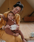 A smiling woman in a mustard yellow dress sits on a wooden floor with a happy baby in brown pajamas, who holds a mushie Bluey x Mushie Snack Cup and an animal-print mug. Another mug rests on the floor nearby.