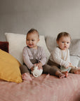 Two babies sit on a couch with light pillows, each holding a sippy cup. One smiles in lavender, the other in white looks serious. Next to them are baby feeding essentials like the mushie Bluey x Mushie Snack Cup and a large yellow cushion.