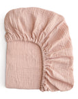 The mushie Extra Soft Muslin Crib Sheet in light pink is folded neatly, showing its crinkled texture and elastic edges. This breathable baby bedding is displayed on a white background.