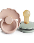 The FRIGG Daisy Natural Rubber Pacifier 2-Pack is shown: one light pink pacifier facing front, and one pale green pacifier with a tan natural rubber nipple lying on its side. Both feature scalloped edges and the FRIGG brand name.