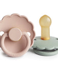The FRIGG Daisy Natural Rubber Pacifier 2-Pack features two scalloped-edge silicone pacifiers: one pale pink daisy with a ring handle and one light green, both embossed with “FRIGG” and made from natural rubber latex.