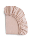 The mushie Stretchy MINI Crib Sheet, shown folded on a white background, features light pink satin fabric with elastic edges and is crafted from breathable organic cotton.