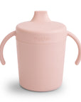 The Mushie Trainer Sippy Cup is a light pink, non-toxic cup with easy-grip handles and a spout lid, featuring the "mushie" label—ideal for young children.
