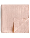 The mushie Organic Cotton Muslin Swaddle Blanket in Blush, featuring a subtle square pattern, is folded with one corner turned up to display its soft organic cotton fabric on a white background.