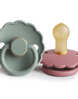 The FRIGG Daisy Natural Rubber Pacifier 2-Pack by FRIGG features two daisy-shaped pacifiers—one in sage green with a forward-facing ring, and one in dusty pink with a natural rubber latex nipple facing up. Both are smooth and have scalloped edges.