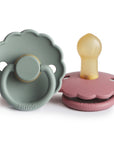 The FRIGG Daisy Natural Rubber Pacifier 2-Pack by FRIGG features two daisy-shaped pacifiers—one in sage green with a forward-facing ring, and one in dusty pink with a natural rubber latex nipple facing up. Both are smooth and have scalloped edges.