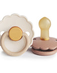 The FRIGG Daisy Natural Rubber Pacifier 2-Pack by FRIGG features two daisy-shaped pacifiers: one upright with a beige front and yellow handle, the other on its side showing a brown base and natural rubber nipple.