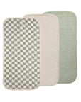 Three Mushie Changing Pad Liners overlap: one features a green and white checkerboard organic cotton design, the second is solid cream, and the third is solid sage green. All have rounded corners and quilted stitching.