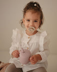 A young child smiles holding a mauve sippy cup while using a FRIGG Natural Rubber 6-Pack pacifier. They wear a white ruffled shirt and grey pants, sitting on a light surface with a neutral background.