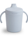 The Mushie Trainer Sippy Cup in light gray features easy-grip handles, a spout lid, and “mushie” embossed on the front. This non-toxic cup helps young children learn to drink independently.