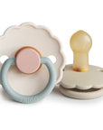 The FRIGG Daisy Natural Rubber Pacifier 2-Pack features two beige, daisy-shaped pacifiers with scalloped edges; one has a blue handle and pink center, the other a yellow natural rubber latex nipple. Both display the FRIGG brand.