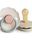 The FRIGG Daisy Natural Rubber Pacifier 2-Pack by FRIGG is shown in beige with scalloped edges; one pacifier displays a blue-gray ring and pink center, while the other reveals a tan natural rubber latex nipple from the side.