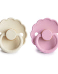 The FRIGG Daisy Natural Rubber Pacifier 2-Pack includes two daisy-shaped pacifiers with scalloped edges, in cream and light pink, round handles with the FRIGG logo, and natural rubber latex nipples for comfort.