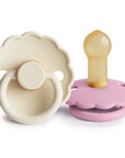 The FRIGG Daisy Natural Rubber Pacifier 2-Pack by FRIGG is shown with a cream daisy-shaped pacifier standing upright and a pink one lying on its side, displaying the natural rubber nipple and scalloped edge.
