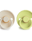 The Mushie FRIGG Rope Natural Rubber Pacifier 2-Pack includes two round, minimalistic pacifiers with textured edges and handles—one off-white, one light green—each featuring a natural rubber latex nipple and “FRIGG” imprinted below.