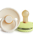 Two baby essentials: Mushie FRIGG Rope Natural Rubber Pacifier 2-Pack—one in pale cream with a white handle, the other in light green with a tan natural rubber latex nipple—are shown from various angles on a white background.