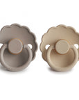 The FRIGG Daisy Natural Rubber Pacifier 2-Pack includes two daisy-shaped pacifiers with scalloped edges in light gray and beige, each featuring natural rubber latex nipples, round handles, and the FRIGG brand name on the front.