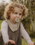 A young child with curly hair looks up while outdoors, using a FRIGG Natural Rubber Pacifier from the 4-Pack. The child wears a light long-sleeve shirt, gray vest, and beige bib, with trees and greenery in the blurred background.