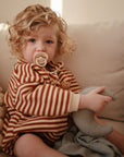 A young child with curly blonde hair sits on a sofa, wearing a red and beige striped outfit and using a FRIGG Natural Rubber Pacifier 4-Pack by FRIGG. The child holds a crescent moon-shaped toy and looks toward the camera.