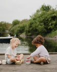 Two young children play with mushie's Bath Boats on a wooden dock by the water. Green trees line the riverbank, and another boat is seen nearby—creating a peaceful scene perfect for outdoor water play.