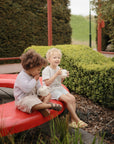Two toddlers sit on a curved red bench in a garden, smiling with mugs in hand. Bushes surround them as their mushie Snack Cup keeps treats secure and spill-proof, making this peaceful outdoor moment even sweeter.
