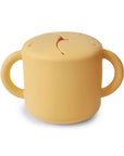 The mushie Snack Cup in Daffodil is a spill-proof toddler snack container with two handles and a flexible, slotted lid that lets little hands reach snacks while preventing spills. Photographed against a white background.