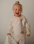 A smiling blonde child in a cream outfit holds a white blanket. A FRIGG Daisy Night Natural Rubber Pacifier from FRIGG, attached with a glow-in-the-dark wooden bead clip, is on their sweater. The background is plain and light-colored.