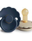 The FRIGG Daisy Natural Rubber Pacifier 2-Pack features two daisy-shaped pacifiers—one dark blue shown facing forward and one beige on its side revealing the natural rubber latex nipple.